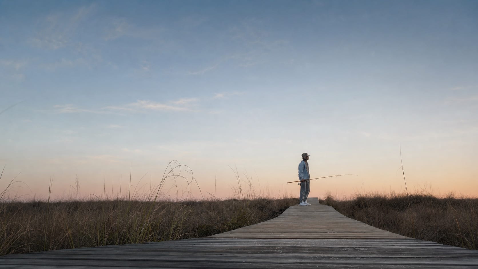 A fisherman standing at the end of a marsh boardwalk at dusk
